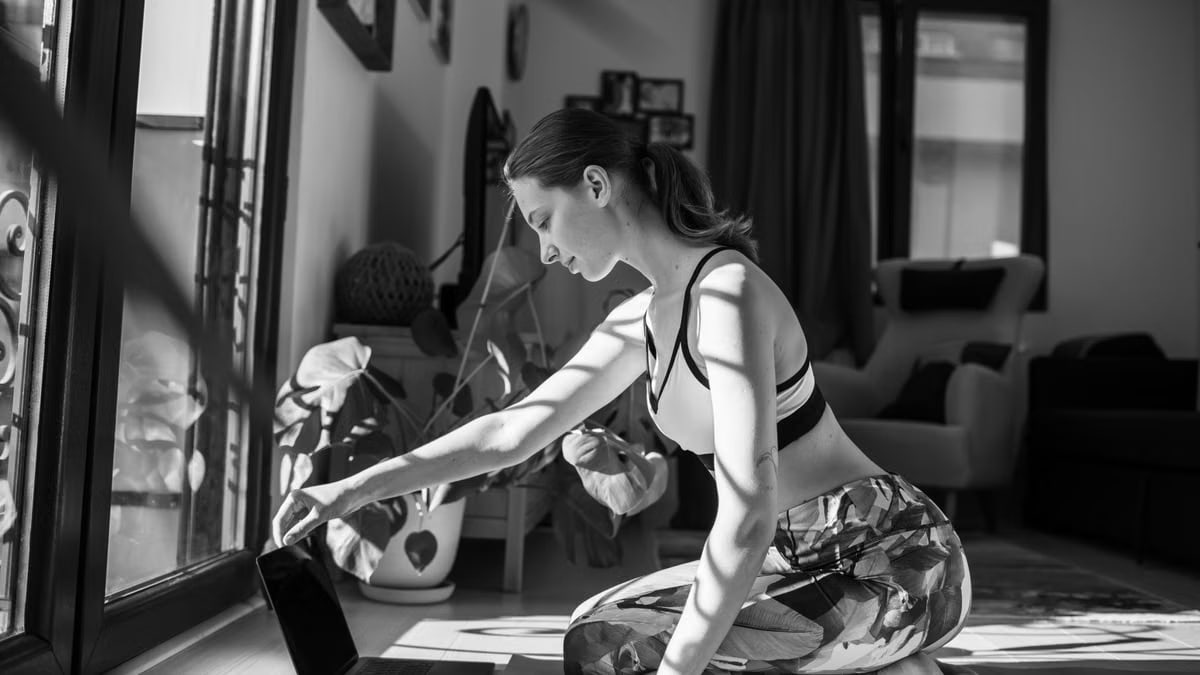 Woman in sports bra using laptop for yoga practice indoors. Sunlight streams through window.